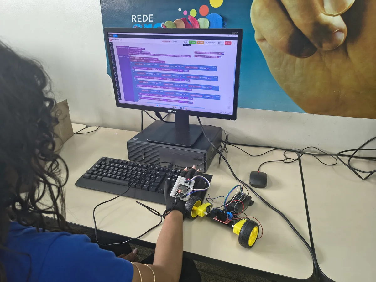 Estudante diante do editor de blocos do Bloquin, com um robô de rodas e uma placa na bancada