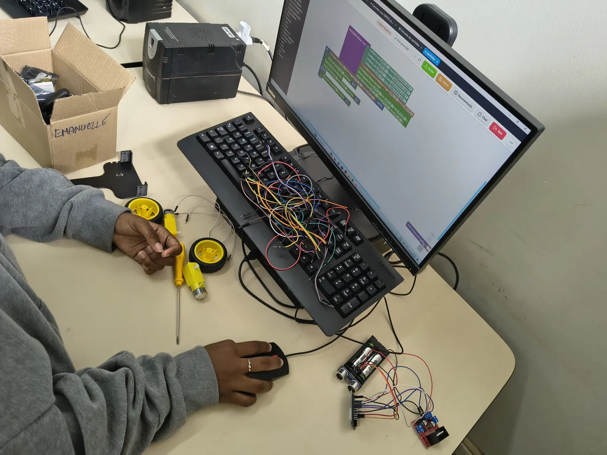 Estudante manuseando um robô de rodas junto a circuitos eletrônicos, com o Bloquin aberto no computador